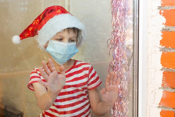 A child in a santa claus hat and a medical mask looks sad from behind the glass, sticking his hands to him. The child inside the building behind the glass. new year in reality 2020. — Fotografia de Stock