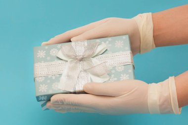 Tıbbi eldivenli eller mavi arka planda mavi bir hediye kutusu tutuyor. Noel salgını. Covid-19 'dan koruma. Sürpriz bir hediye. Kendine dikkat et. Yeni Yıl 2021.