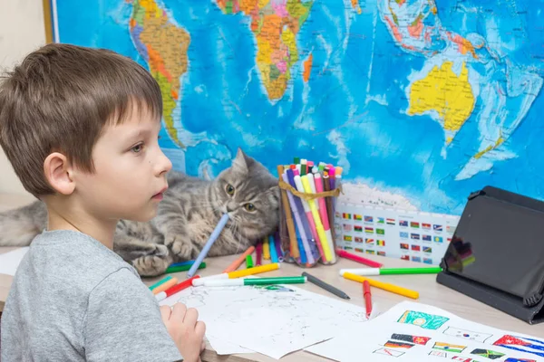 the child sits thoughtfully at the table against the background of the geographical map. Nearby, a cat gnaws at a felt-tip pen. soft focus The idea is to educate children during a pandemic