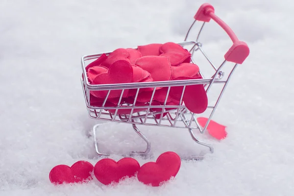 Der Warenkorb ist mit kleinen Herzen gefüllt. Der Wagen steht im Schnee, drei Herzen sind in der Nähe. Nahaufnahme, horizontales Foto. Idee - Shopping für Liebste am Valentinstag. — Stockfoto