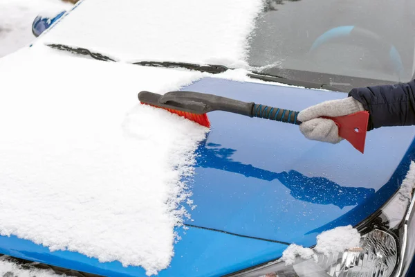 A human hand in a glove cleans a blue car from snow with a special brush. A hand sweeps snow from the hood of the car. Horizontal photo. the consequences of a blizzard.