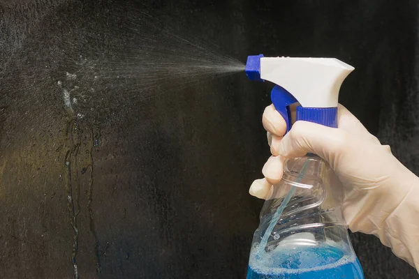 Zbliżenie ręki trzymającej butelkę niebieskiego detergentu rozpyla chemikalia na ciemną powierzchnię. Koncepcja - czyszczenie, dezynfekcja. Utrzymanie czystości w domu, ogólne sprzątanie. — Zdjęcie stockowe
