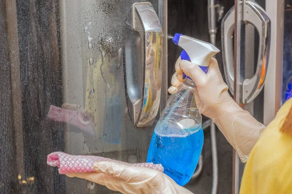 在女人的手里 瓶子里有一种蓝色的液体清洁剂和一件破衣服 把产品喷在肮脏的玻璃门上 清洁公寓 浴室的概念 概念是清洁和秩序 图库图片