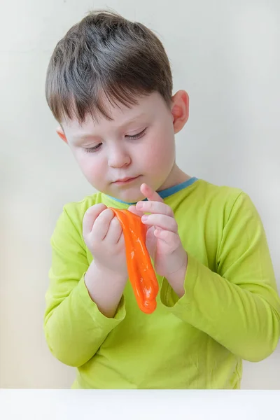 Een jongen met een Europees uiterlijk speelt met oranje slijm. Het kind lacht niet, hij kijkt naar iets. Het concept is tastbare sensaties, de ontwikkeling van motoriek bij kinderen. — Stockfoto