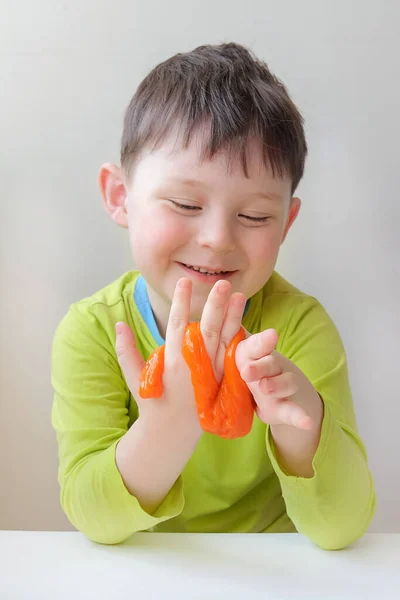 Jongen in groene kleren speelt met oranje slijm. Het kind glimlacht, kijkt hoe het slijm aan zijn vingers kleeft. Verticaal. Het concept is educatieve spelletjes voor kinderen, de ontwikkeling van creatief denken. — Stockfoto