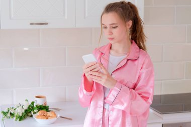 Pembe ipek pijamalı bir kız elinde telefonla mutfakta duruyor. Kuryeleri, postaları kontrol eder. Günaydın. Masada bir kupa, taze kruvasanlar ve beyaz taze çiçekler var..