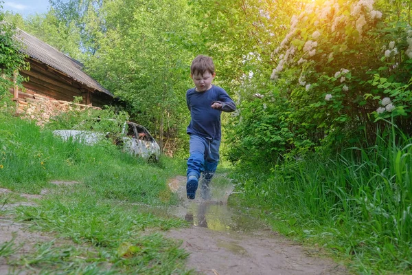 Ребенок бежит через лужи в сапогах. Забавный, эмоциональный мальчик. Брызги воды. Дошкольник среди зелени, на улице, после дождя. Горизонтальное фото. — стоковое фото