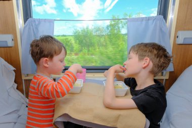 İki kardeş pencerenin yanındaki kompartımanda bir masada oturuyorlar. Hazır makarna yiyorlar. Öğle yemeği ya da kahvaltı. Keyifle seyahat et. Yatay fotoğraf