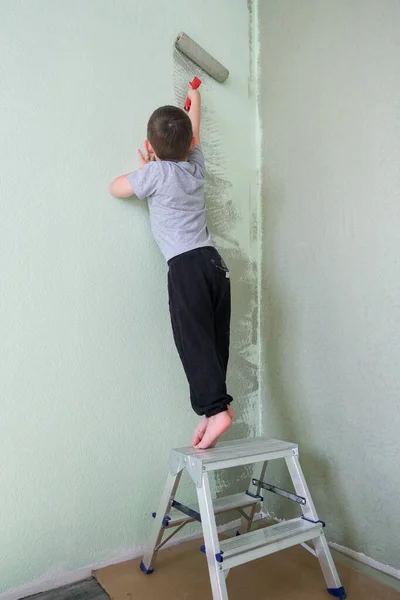 a child stands on the stairs with his back to the camera. A boy in a gray T-shirt and black pants. He paints the wall green with a roller. Vertical photo