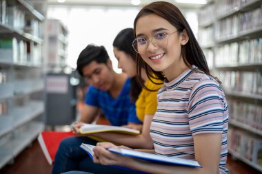Grup Asyalı Öğrenciler gülümseyip kitap okuyor ve dizüstünü kütüphanede ödev tartışan öğrenciler için kullanıyor.
