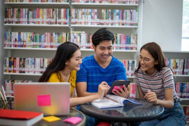Grup Asyalı Öğrenciler gülümseyip kitap okuyor ve dizüstünü kütüphanede ödev tartışan öğrenciler için kullanıyor.
