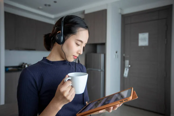 ヘッドフォンを着用し デジタルタブレットビデオを使用してアジアの女性会議や自宅のキッチンでコーヒーを飲みます — ストック写真