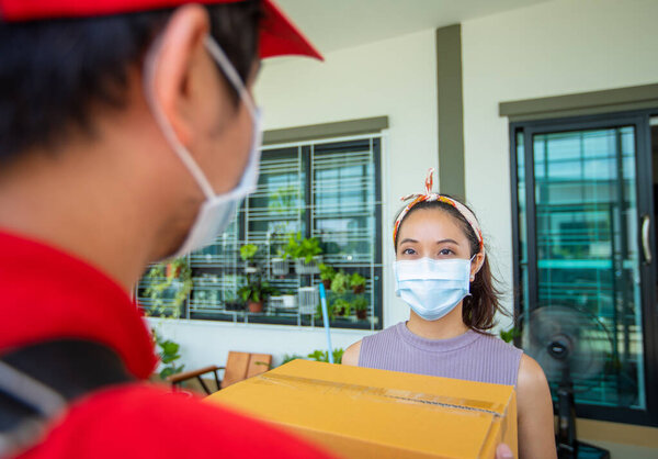Asian delivery servicemen wearing a red uniform with a red cap and face mask handling cardboard boxes to give to the female customer in front of the house. Online shopping and Express delivery 