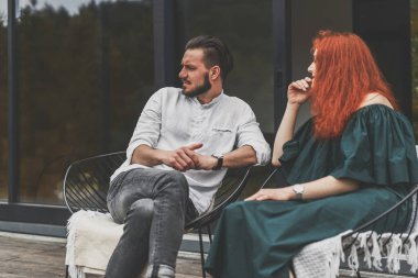 Ahşap verandada oturan ve arka planda rahatlayan şık bir çift.