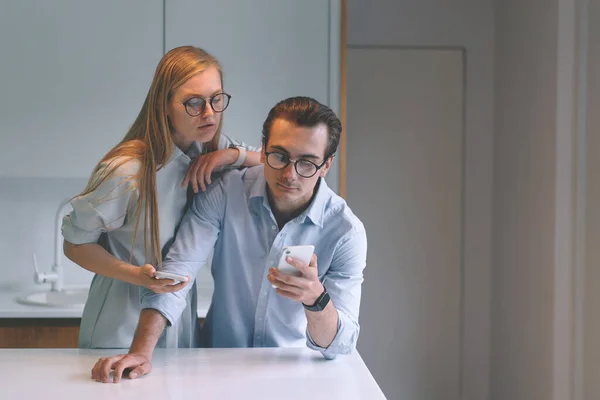 Vista frontal de la pareja joven usando teléfonos inteligentes mientras está de pie en la cocina —  Fotos de Stock