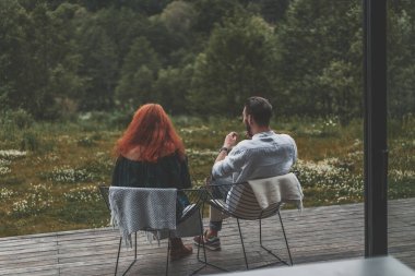 Genç bir çift modern evin terasında oturuyor, güzel manzaranın tadını çıkarıyorlar.