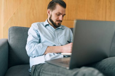 Sinirli esmer adam dizüstü bilgisayarla çalışıyor ve notlar alıyor.