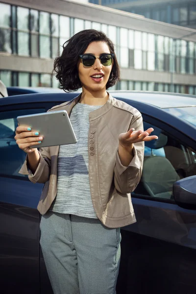Unga leende kvinna i solglasögon med tablett står nära bilen — Stockfoto