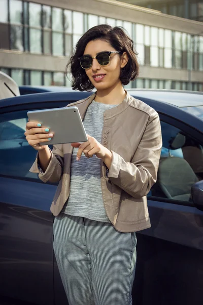 Unga leende kvinna i solglasögon med tablett står nära bilen — Stockfoto