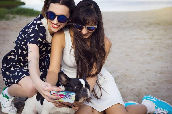 Deux belles jeunes femmes sur la plage prenant des photos selfie avec des bulldogs français — Photo