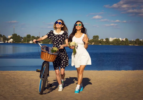 Twee mooie meisjes op het strand met fiets en bloemen in mand — Stockfoto