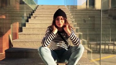 Kot pantolon ve çizgili yelek giyen beyaz bir kadının merdivenlerde oturduğu yavaş çekim videosu.