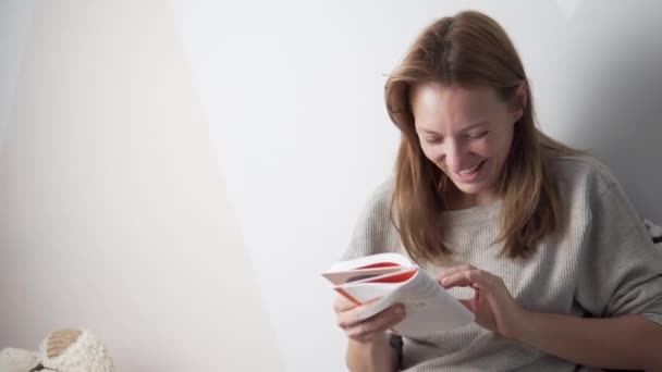 Een vrouw leest een grappig boek thuis op de bank — Stockvideo