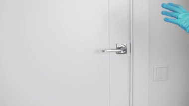 Ağır çekimde, tıbbi eldiven giymiş bir kadın kapıyı kolunun arkasından açar.