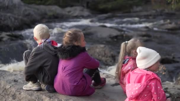 Crianças sentam-se em uma rocha e olham para um rio de montanha durante uma caminhada em família — Vídeo de Stock