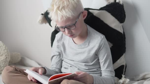 A blond boy with glasses reads a book at home on the bed — Stock Video