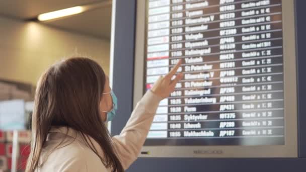 Žena v lékařské masce stojí na letišti u odletové tabule během pandemie covid-19. — Stock video