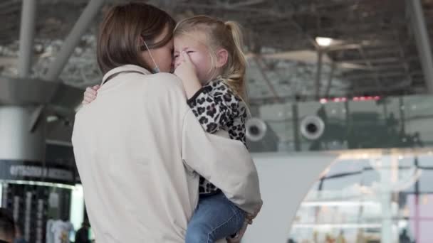 Una madre con una máscara médica sostiene a una hija llorando en sus brazos en el aeropuerto en el área de salida — Vídeos de Stock