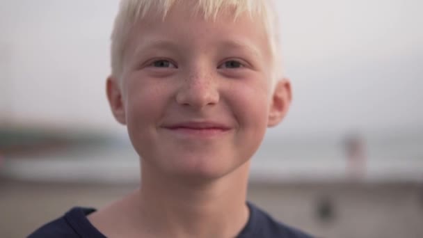 Крупним планом красивий альбіносів хлопчик показує язик на тлі розмитого моря — стокове відео