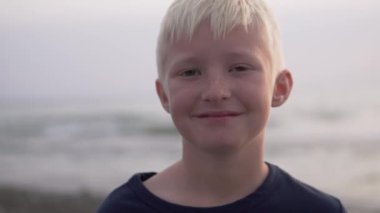 Yakın planda kalan sarışın çocuk gün batımında denize gülümsüyor.