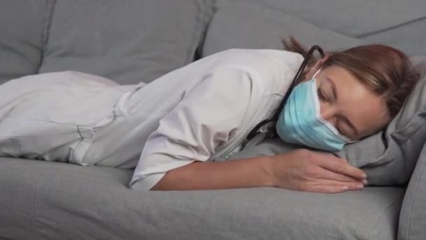 A woman doctor in a medical mask falls on the sofa without strength and falls asleep after a hard shift in the hospital — Stock Video