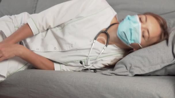 Exhausted or bored young sleepy woman doctor in a medical gown and mask falls down on sofa — Stock Video