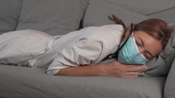 A tired and sleepy woman doctor in a medical mask falls on the couch and falls asleep. In a dream — Stock Video