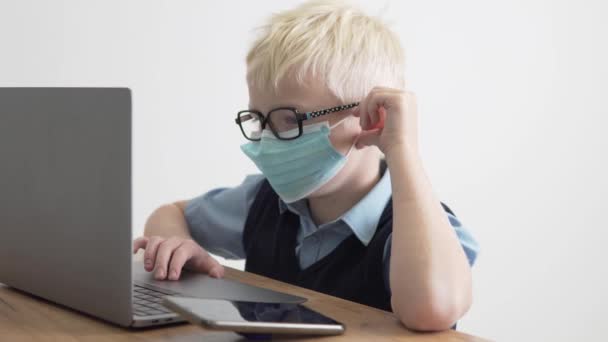 Ένα αγόρι σχολιαρόπαιδο με γυαλιά και μια ιατρική μάσκα στο πρόσωπό του ασχολείται με το σχολείο σε έναν υπολογιστή — Αρχείο Βίντεο