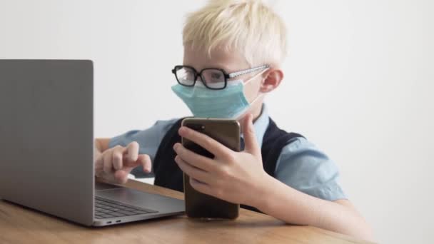 Un niño colegial con una máscara médica en la cara haciendo clases en casa en una computadora — Vídeos de Stock