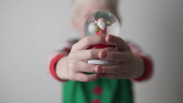 Schöner blonder Junge hält eine Schneekugel mit Weihnachtsmann und Schnee in die Weihnachtsfeiertage — Stockvideo