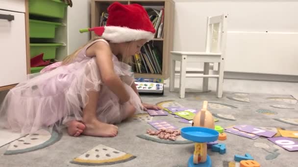 Una niña inteligente con un sombrero de Santa Claus aprende a contar usando un juguete de balanzas y pesas con números — Vídeo de stock