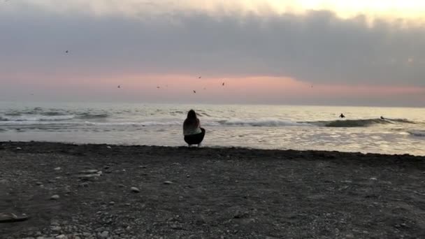 Una mujer mira la puesta de sol en el mar y se burla de volar sobre el agua en el otoño en Sochi en una playa de guijarros — Vídeos de Stock