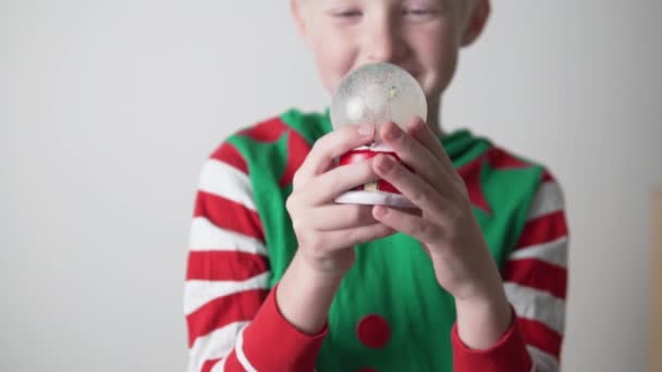 Seorang anak laki-laki berpakaian seperti gnome memegang bola kaca salju dengan Santa Claus di tangannya pada hari libur Natal — Stok Video
