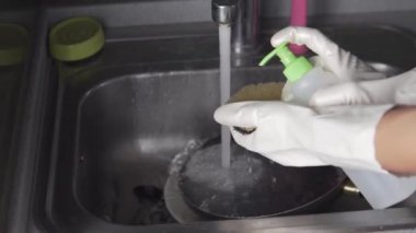 Koruyucu eldivenli bir kadın deterjanı süngere sıkıyor ve kirli bulaşıkları lavaboda yıkıyor.