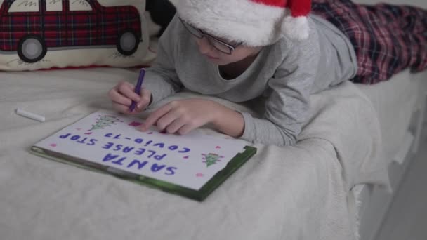A boy in a Santa Claus cap writes on paper his New Years wish — Stock Video
