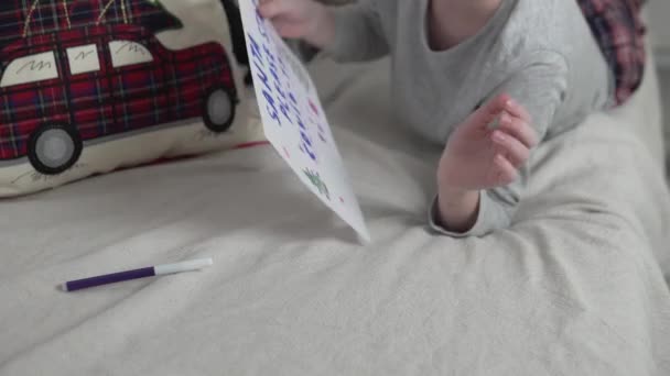Un niño con una gorra de Navidad dobla una carta para Papá Noel en casa en la cama — Vídeo de stock