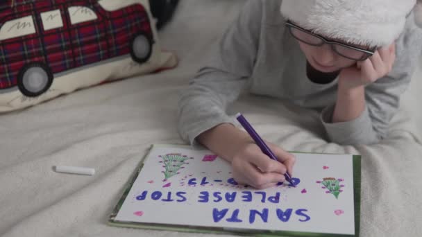 En söt pojke med glasögon och en röd mössa skriver ett brev till jultomten med önskan att stoppa Covid-19 — Stockvideo