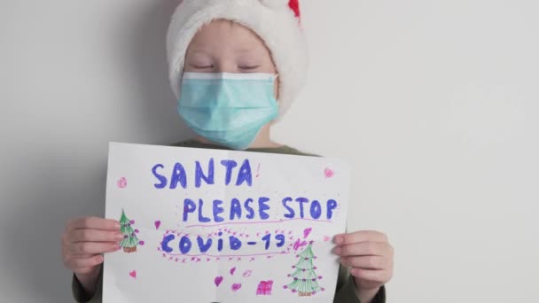 Seorang anak bertopi Santa Claus dan topeng medis di wajahnya berdiri dengan poster bertuliskan — Stok Video