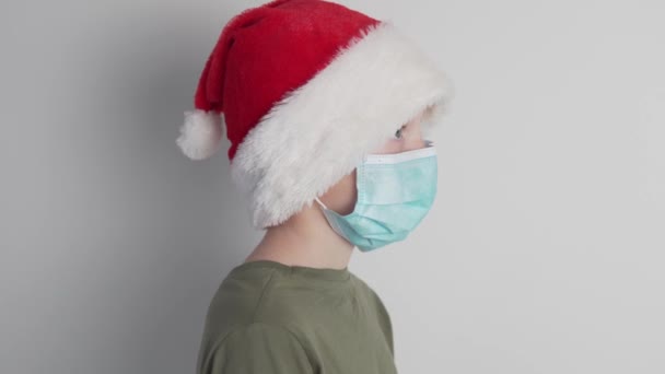Seorang anak laki-laki bertopeng medis di wajahnya dan mengenakan topi Santa Claus berdiri diprofilkan ke kamera, — Stok Video