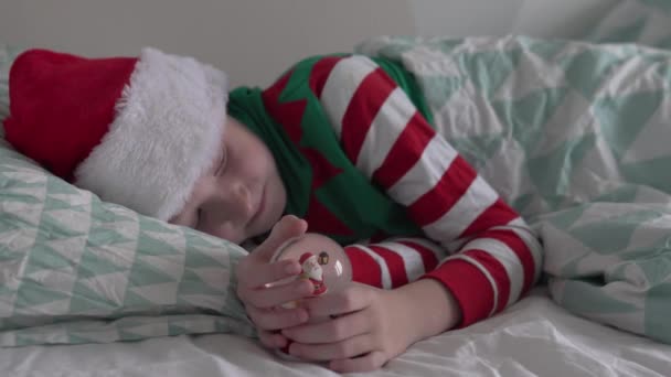 圣诞节早上，一个穿着圣诞老人帽和侏儒睡衣的可爱男孩在床上醒来 — 图库视频影像
